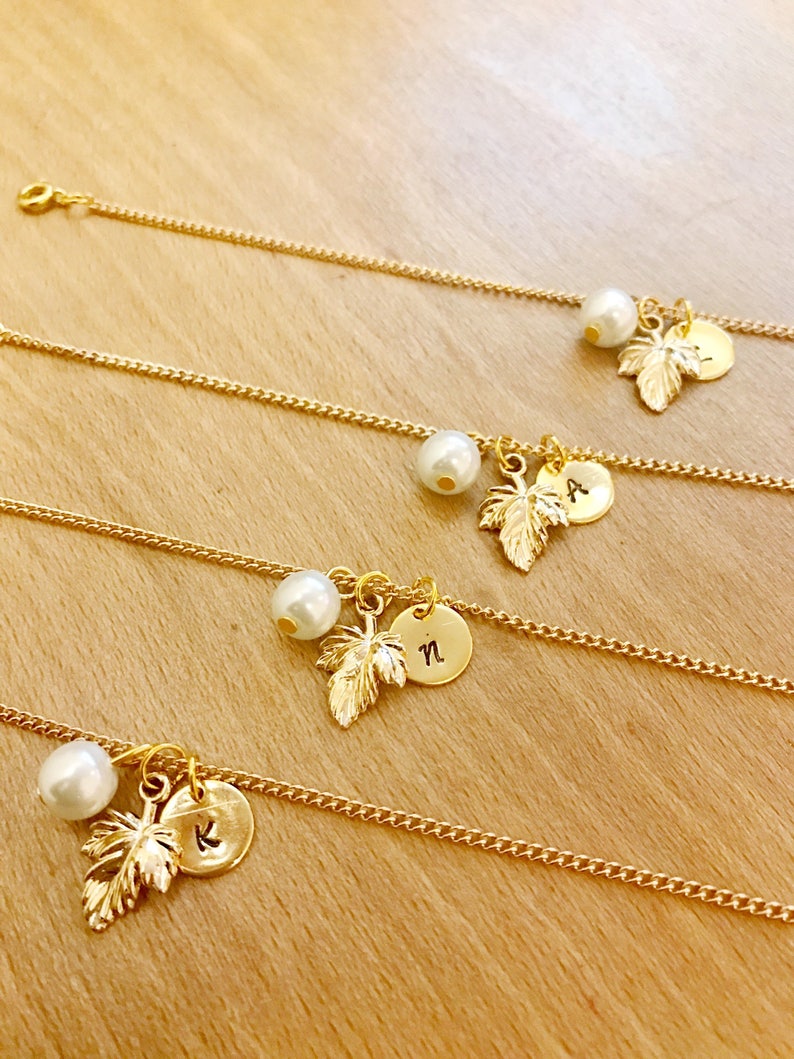 May include: Four gold chain bracelets with a pearl and a gold leaf charm with a letter on a round disc. The letters are K, N, A, and a fourth letter that is not visible.