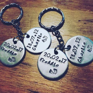 Peut inclure: Trois porte-cl&eacute;s en argent grav&eacute;s avec des dates et des noms. Les porte-cl&eacute;s portent les noms "Freddie" et "Charlie" avec leurs dates de naissance respectives. Chaque porte-cl&eacute;s a un petit symbole de pied.