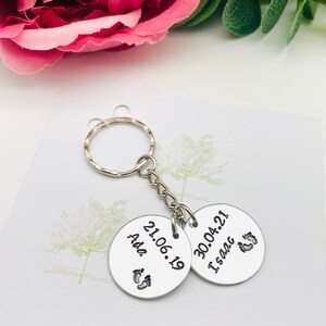 Peut inclure: Deux porte-cl&eacute;s en argent grav&eacute;s avec des dates et des noms. Le porte-cl&eacute;s de gauche indique "Ada 21.06.19" et comporte un symbole de pied. Le porte-cl&eacute;s de droite indique "30.04.21 Isaac" et comporte un symbole de pied.