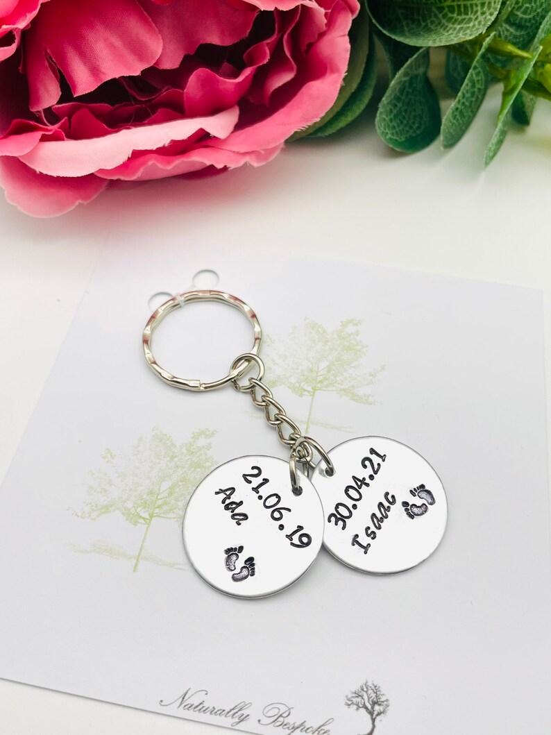Peut inclure: Deux porte-cl&eacute;s en argent grav&eacute;s avec des dates et des noms. Le porte-cl&eacute;s de gauche indique "Ada 21.06.19" et comporte un symbole de pied. Le porte-cl&eacute;s de droite indique "30.04.21 Isaac" et comporte un symbole de pied.