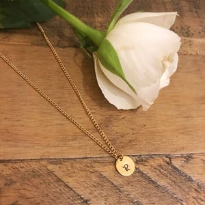 May include: A gold chain necklace with a small round pendant featuring the letter "R" engraved on it. The necklace is lying on a wooden surface next to a white rose.