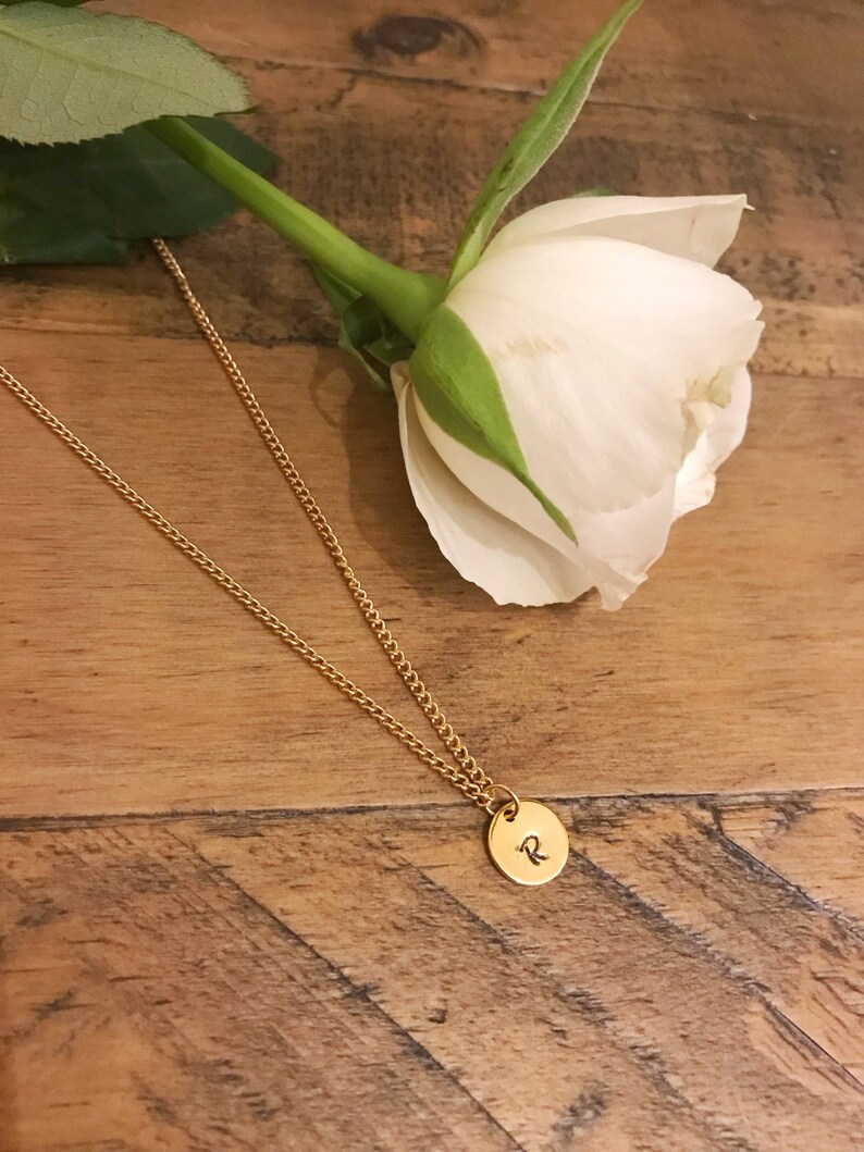 May include: A gold chain necklace with a small round pendant featuring the letter "R" engraved on it. The necklace is lying on a wooden surface next to a white rose.
