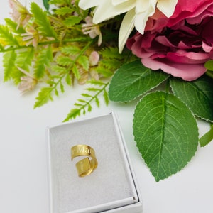 May include: A gold-colored ring with the word "Mar" engraved on it. The ring is in a white box, surrounded by pink and white flowers.