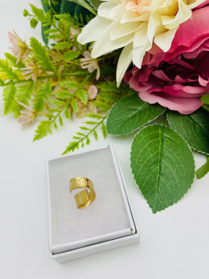 May include: A gold-colored ring with the word "Mar" engraved on it. The ring is in a white box, surrounded by pink and white flowers.