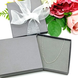 May include: A silver chain necklace in a gray gift box with a white satin ribbon. The box is surrounded by pink and white flowers.