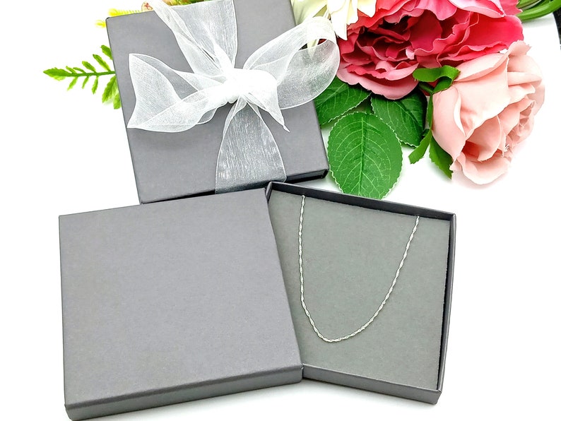 May include: A silver chain necklace in a gray gift box with a white satin ribbon. The box is surrounded by pink and white flowers.