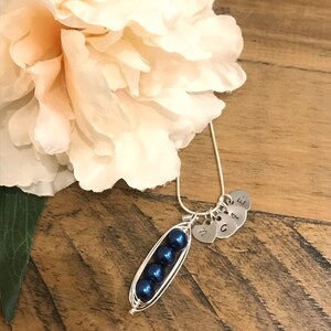May include: A silver necklace with a pea pod charm made of wire and blue pearls. The charm has four small silver hearts with the letters M, G, A, and E attached.