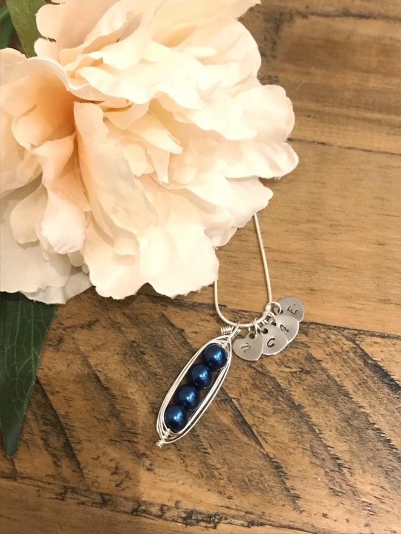 May include: A silver necklace with a pea pod charm made of wire and blue pearls. The charm has four small silver hearts with the letters M, G, A, and E attached.