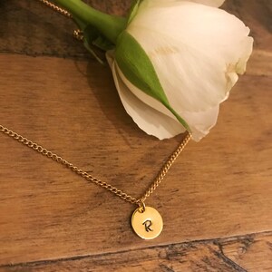 May include: A gold chain necklace with a small round pendant featuring the letter "R" engraved on it. The necklace is lying on a wooden surface with a white rose in the background.
