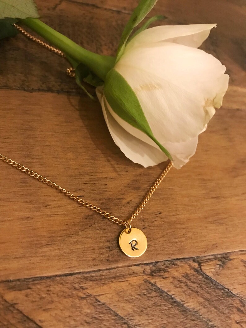 May include: A gold chain necklace with a small round pendant featuring the letter "R" engraved on it. The necklace is lying on a wooden surface with a white rose in the background.