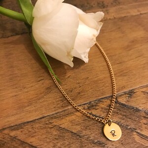 May include: A gold chain necklace with a small round pendant featuring the letter "R" engraved on it. The necklace is lying on a wooden surface next to a white rose.