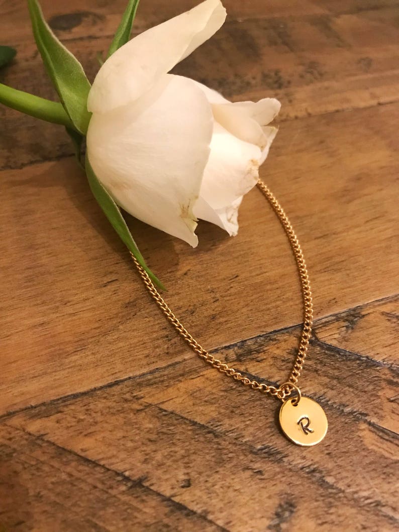 May include: A gold chain necklace with a small round pendant featuring the letter "R" engraved on it. The necklace is lying on a wooden surface next to a white rose.