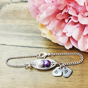 May include: A silver chain bracelet with a charm that resembles a pea pod with two pearls inside. One pearl is white and the other is purple. The bracelet also has two silver heart charms with the letters "D" and "P" engraved on them.