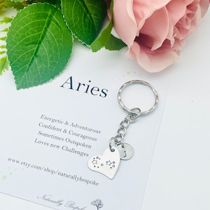 Könnte beinhalten: Ein silberner Schlüsselanhänger mit einem herzförmigen Anhänger, der mit einem Sternmuster und dem Buchstaben "J" verziert ist. Die Karte trägt die Aufschrift "Aries" und listet Eigenschaften auf, die mit dem Sternzeichen verbunden sind, darunter "Energetisch & Abenteuerlustig", "Selbstbewusst & Mutig", "Manchmal offenherzig" und "Liebt neue Herausforderungen". Die Karte enthält auch die Website-Adresse "www.etsy.com/shop/naturallybespoke" und den Text "Naturally Bespoke".