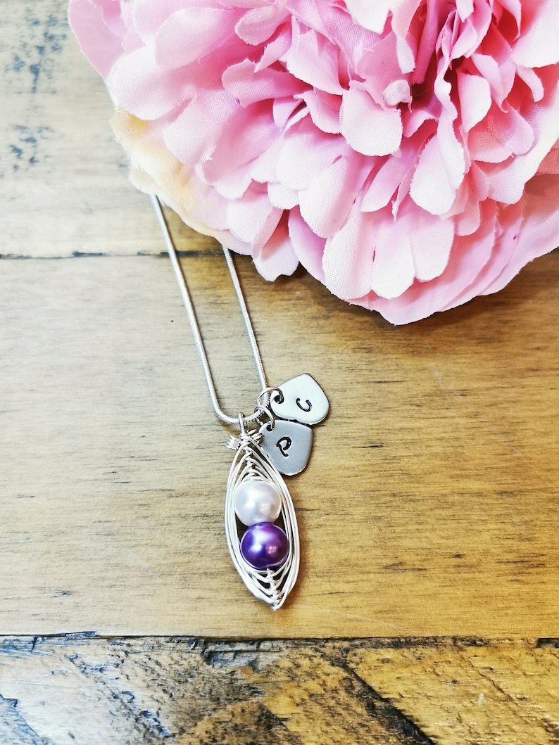 May include: A silver necklace with a pea pod charm containing three pearls. The pearls are white and purple. The necklace also has two silver heart charms with the letters 'P' and 'C' on them.