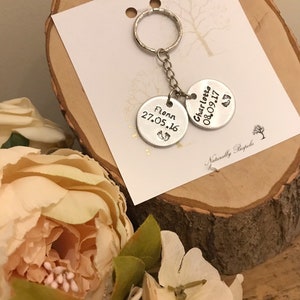 Peut inclure: Porte-cl&eacute;s argent&eacute; avec deux &eacute;tiquettes rondes. L'&eacute;tiquette du haut indique "Charlotte 08.09.17" avec un symbole de pied. L'&eacute;tiquette du bas indique "Flynn 27.05.16" avec un symbole de pied. Le porte-cl&eacute;s est sur une surface en bois avec une carte blanche qui dit "Naturally Bespoke".