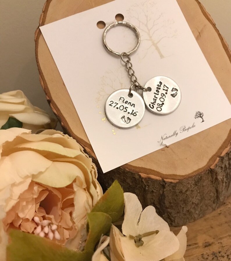 Peut inclure: Porte-cl&eacute;s argent&eacute; avec deux &eacute;tiquettes rondes. L'&eacute;tiquette du haut indique "Charlotte 08.09.17" avec un symbole de pied. L'&eacute;tiquette du bas indique "Flynn 27.05.16" avec un symbole de pied. Le porte-cl&eacute;s est sur une surface en bois avec une carte blanche qui dit "Naturally Bespoke".