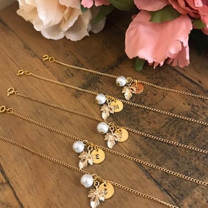 May include: Five gold chain bracelets with a pearl and a gold leaf charm. Each charm has a different letter engraved on it.