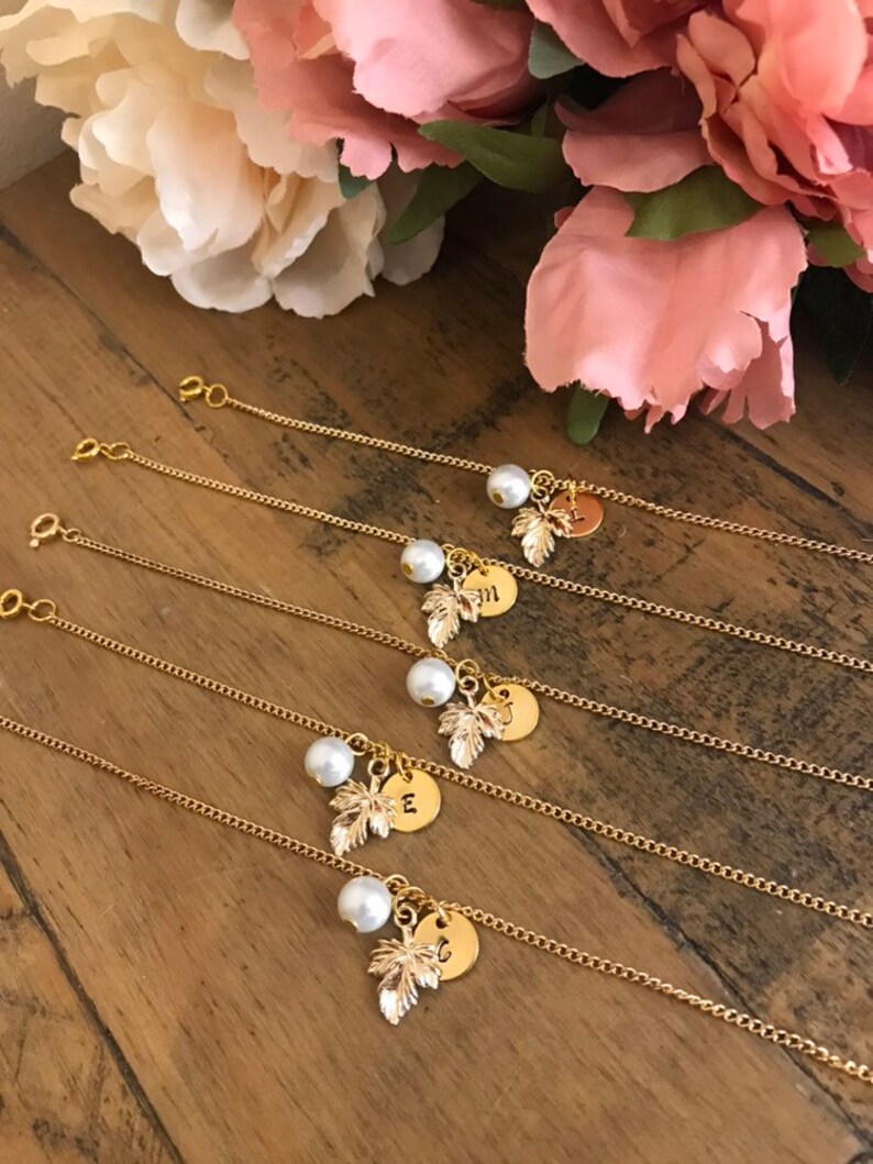 May include: Five gold chain bracelets with a pearl and a gold leaf charm. Each charm has a different letter engraved on it.