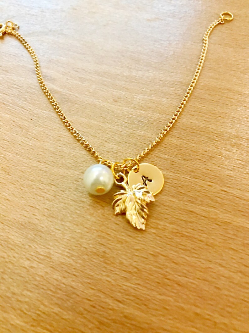 May include: A gold chain bracelet with a pearl charm, a gold leaf charm, and a gold disc charm with the letter "A" engraved on it.