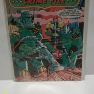 May include: A comic book cover featuring Judge Dredd's Crime File. The cover art depicts Judge Dredd and two other characters in a futuristic setting. The text on the cover reads "Judge Dredd's Crime File" and "Get to work on that sludge. I want to identify this citizen before he dissolves completely."