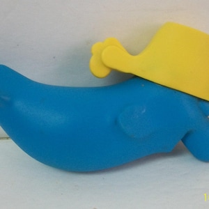 May include: A blue plastic toy shaped like a whale with a yellow hat. The toy has a curved body and a spout-like feature at the front. The yellow hat is positioned on top of the whale's body.