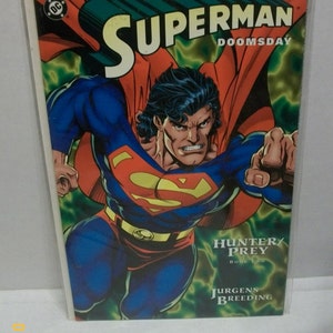 May include: A comic book cover featuring Superman in a red and blue suit, flying through a green and yellow background. The title is "Superman Doomsday: Hunter/Prey Book Two" with the author's name "Jurgens Breeding" below.