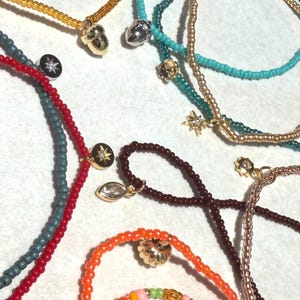 May include: A collection of beaded necklaces and bracelets in various colors including red, orange, teal, dark gray, and gold. The jewelry pieces feature small metallic charms shaped like acorns, stars, and flowers, some with embedded clear stones.