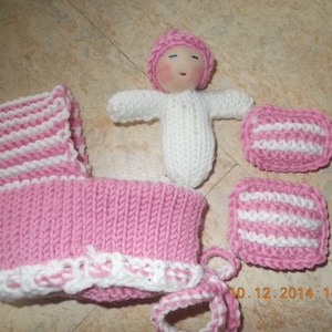Könnte beinhalten: Eine rosa und weiße gestrickte Babypuppe mit einer rosa gestrickten Mütze. Die Puppe trägt ein weißes gestricktes Kleid und hält eine rosa und weiße gestrickte Decke. Die Puppe sitzt auf einer rosa und weißen gestrickten Decke.