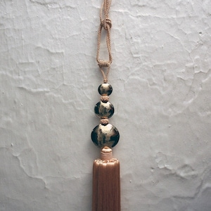 Può includere: Un fiocco beige con un accento di perline decorative dorate e nere. Il fiocco è appeso a una corda beige.