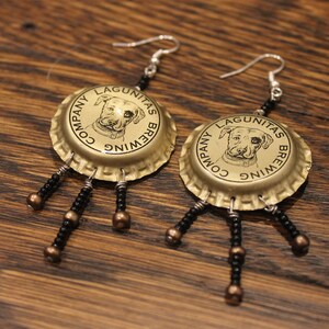 May include: A pair of earrings made from gold bottle caps with black beads hanging from them. The bottle caps have the text "Lagunitas Brewing Company" and a dog's head on them.