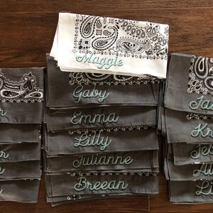 May include: A stack of gray bandanas with white and black paisley designs. Each bandana has a different name embroidered in teal lettering. The names include Maggie, Gaby, Taylor, Jenna, Heather, Lauren, Gabby, Emma, Lilly, Julianne, Breean, Jaime, Krista, Jessy, Alex, and Liz.