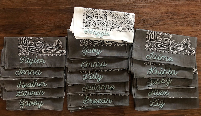 May include: A stack of gray bandanas with white and black paisley designs. Each bandana has a different name embroidered in teal lettering. The names include Maggie, Gaby, Taylor, Jenna, Heather, Lauren, Gabby, Emma, Lilly, Julianne, Breean, Jaime, Krista, Jessy, Alex, and Liz.