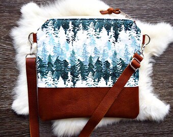 May include: A small crossbody bag with a brown leather strap and a white fabric front featuring a watercolor print of a forest with blue and green trees.