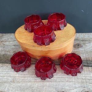 May include: Six ruby red glass votive candle holders with a geometric design. The candle holders are arranged on a wooden, round, lidded box. The background is a dark gray color, and the surface is a light wood.