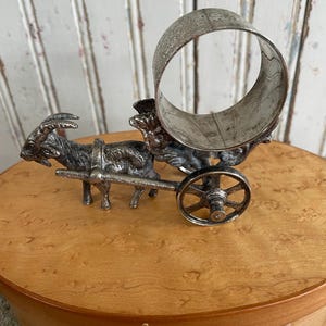 May include: A silver-toned metal napkin ring shaped like a goat pulling a small cart. The goat has detailed horns and is harnessed to the cart, which features a large ring for holding napkins. The piece is set on a wooden surface.