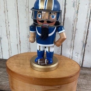 May include: A wooden nutcracker figurine dressed in a blue and gold football uniform with a matching helmet and face mask. The figure has a black mustache and beard, stands on a gold circular base, and holds a small brown football at its feet.