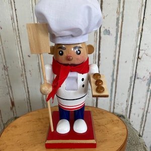 May include: A wooden chef nutcracker figurine, wearing a white chef's hat, red scarf, and blue pants. The figurine holds a wooden paddle and a tray of baked goods. The base is red, and the figurine is approximately 10 inches tall.