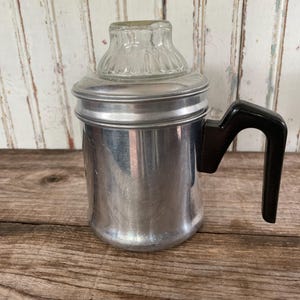 May include: A vintage aluminum percolator coffee pot featuring a cylindrical body, a black plastic side handle, and a clear glass knob on the lid. The metallic surface shows a polished finish with subtle signs of wear and light surface scratches.