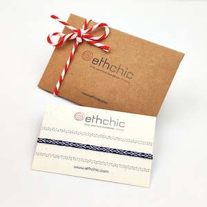 以下が含まれることがあります： 「ethchic」のロゴと「latin american handmade - france」と「www.ethchic.com」のテキストが書かれた2枚のカード。カードは白い表面の上に置かれています。1枚のカードには、青と白の紐のブレスレットが置かれています。