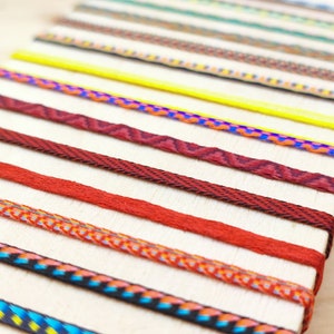 Pode incluir: Um close-up de uma variedade de cordões ou fitas tecidas coloridas dispostas em filas sobre uma superfície de madeira clara. Os cordões são de diferentes cores, padrões e texturas.