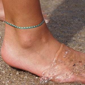 Peut inclure: Gros plan sur un pied nu de femme avec un bracelet de cheville vert et blanc à son cheville. Le pied est debout sur une plage de sable.