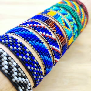Può includere: Una pila di braccialetti colorati con perline blu, bianche, rosse, gialle, verdi e arancioni. I braccialetti sono realizzati con piccole perline rotonde e sono intrecciati insieme in un motivo.
