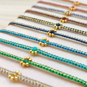 Puede incluir: Primer plano de varias pulseras coloridas. Cada pulsera presenta un cordón tejido en varios colores, incluyendo verde, azul y rojo, con detalles dorados. Un pequeño dije en forma de flor con un centro de piedras preciosas adorna cada pulsera.