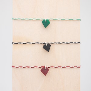 Può includere: Tre braccialetti con ciondoli di diversi colori. Il braccialetto superiore ha un ciondolo verde, il braccialetto centrale ha un ciondolo nero e il braccialetto inferiore ha un ciondolo rosso.