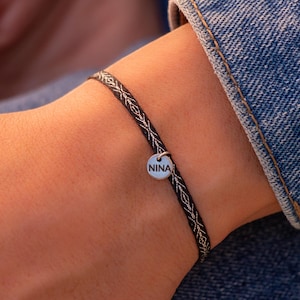 May include: A braided bracelet in black and silver, featuring a small, circular charm inscribed with the name "NINA". The bracelet is displayed on a wrist, with a denim jacket sleeve visible in the background.