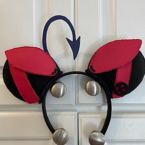 Op de afbeelding: Een zwart hoofdband met rode en zwarte oren en een blauwe staart. De oren hebben een zwart-rood streepdessin en een zwarte knop met een witte X.