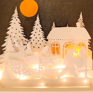 Può includere: Una scena natalizia ritagliata in carta bianca con una casa, alberi, una slitta, renne e Babbo Natale. La scena è illuminata da luci bianche calde e una luna gialla.
