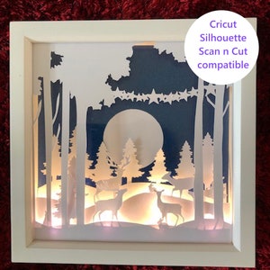 May include: A white framed shadow box with a paper cut design of a moonlit forest scene with deer. The design is illuminated from behind with warm white LED lights. The box is labelled "Cricut Silhouette Scan n Cut compatible".