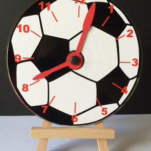 Puede incluir: Un reloj de fútbol negro y blanco con manecillas rojas, mostrado en un pequeño caballete de madera.